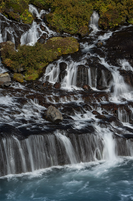 A steep basalt column canyon in Iceland with wet, amber-toned rocks in the foreground and a calm turquoise river cutting between towering rows of vertical, geometric stone pillars under a soft sky.
