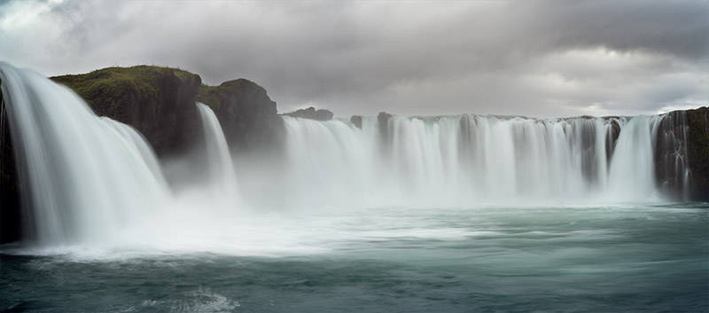 A wide, panoramic long-exposure photograph of Goðafoss waterfall in Iceland under a dark, moody storm sky. Multiple silky white curtains of water cascade over a curved horseshoe cliff of dark basalt rocks into a vibrant, flowing aqua-blue river basin below.