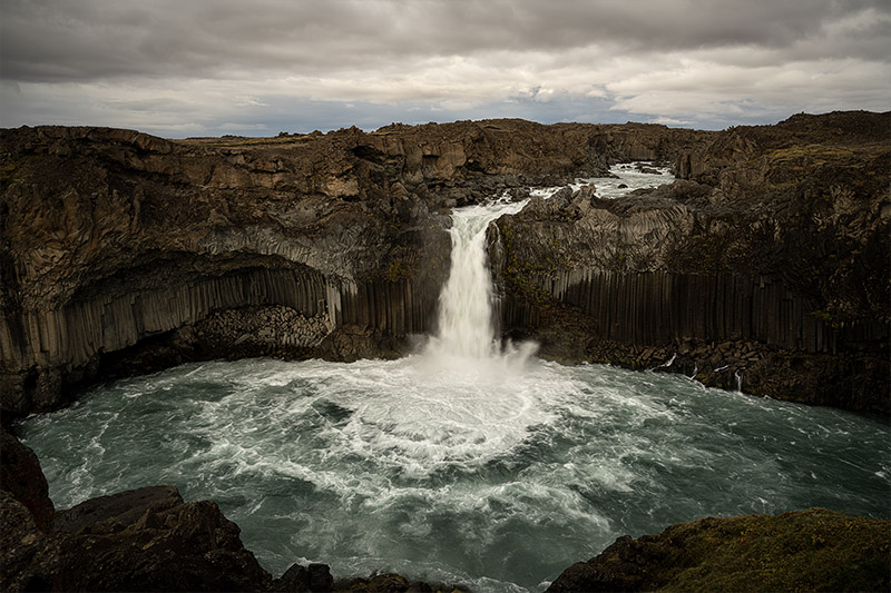  powerful white waterfall plunging into a large, frothy basin surrounded by dark, symmetric basalt column cliffs under a cloudy sky in Iceland.