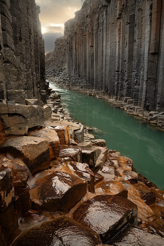 A steep basalt column canyon in Iceland with wet, amber-toned rocks in the foreground and a calm turquoise river cutting between towering rows of vertical, geometric stone pillars under a soft sky.